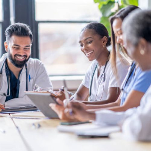 A small group of four medical professionals sit around a boardroom table as they meet to discuss patient cases.  They are each dressed professionally in scrubs and lab coats as they focus on working together.